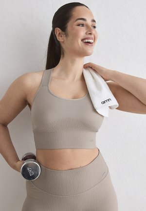 Frau in beiger Sportkleidung hält eine 4-kg-Hantel und ein weißes Handtuch über der Schulter, lächelt und blickt zur Seite.