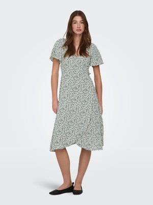 Kvinde stående iført en grøn midi-kjole med hvidt blomsterprint, korte flagrende ærmer og sorte flade sko på en ensfarvet lys baggrund.