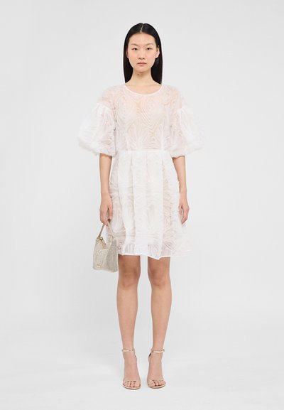 Robe courte blanche avec des motifs floraux texturés, des manches bouffantes et une taille cintrée. Associée à un sac à main métallique et des sandales dorées à lanières.