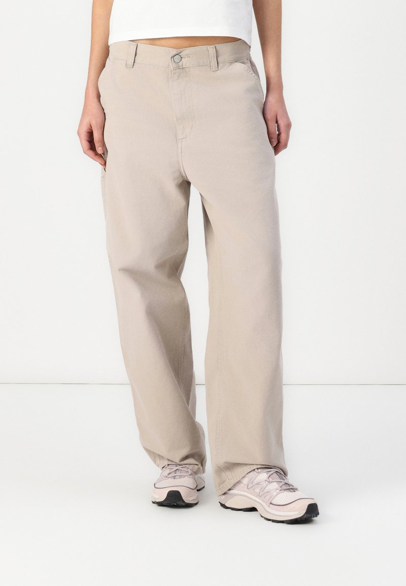 Pantaloni di un beige chiaro con una vestibilità dritta, realizzati in cotone. Presentano tasche frontali, una zip e una chiusura con bottone. Indossati con sneakers chiare.