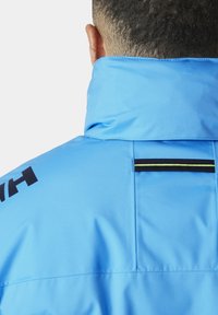 Lichtblauwe jas met hoge kraag, voorzien van zwarte tekst en een horizontale stripe in zwart en geel op de rug. Glad textuur.
