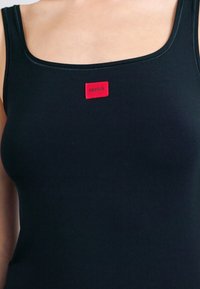 Μαύρο τοπάκι με τετράγωνο ντεκολτέ, εφαρμοστή σχεδίαση και έντονη κόκκινη ορθογώνια ετικέτα με την επιγραφή "HUGO" στο μπροστινό μέρος.