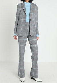 Siwy garnitur w kratę z jasnoniebieskimi akcentami, składający się z dopasowanej marynarki, prostych spodni i prążkowanego niebieskiego swetra. Białe, spiczaste botki na kostkę.