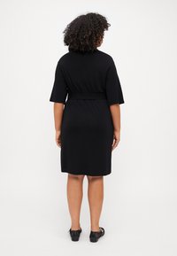 Černé šaty ke kolenům s krátkými rukávy, opaskem v pase a hladkým povrchem. Nosené s černými plochými botami, pohled zezadu.