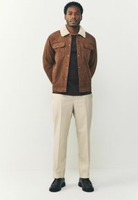 Geacă din suede maro cu guler alb din blană artificială, cămașă neagră dedesubt, pantaloni bej deschis și pantofi negri masivi. Design simplificat și curat.