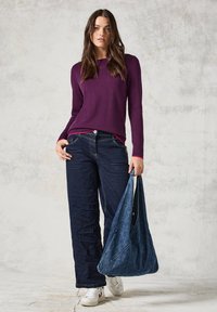 Lila langärmliger Pullover kombiniert mit dunkelblauen, weit geschnittenen Jeans. Eine Denim-Schultertasche wird in der rechten Hand gehalten. Weiße Sneakers runden den Look ab.