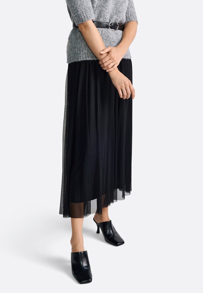 Zwarte geplooide maxi rok met een doorschijnende onderlaag, gecombineerd met een grijze gebreide top en een zwarte riem. G gedragen met zwarte high heel muilen.