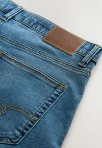 Jean en denim bleu avec poche arrière, couture jaune et patch en cuir marron marqué "DENIM SUPPLY."