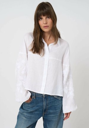 Femme aux longs cheveux bruns portant une chemise blanche ample à boutons avec une broderie florale sur les manches et un jean bleu, les mains dans les poches.