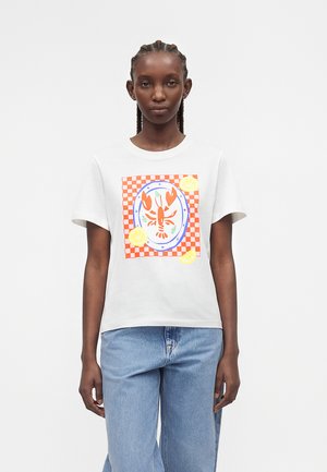 Ung kvinde iført en hvid t-shirt med en rød hummergrafik på en ternet orange og hvid baggrund, kombineret med lyseblå jeans.