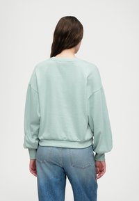 Mentazöld, oversized kapucnis pulóver, leengedett vállakkal, bordás mandzsettákkal és aljjal. Simás textúra és laza szabás. Világoskék farmernadrággal viselve.