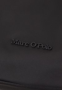 Sac en tissu noir avec une surface texturée, arborant un logo "Marc O'Polo" en relief, brillant et mis en avant.