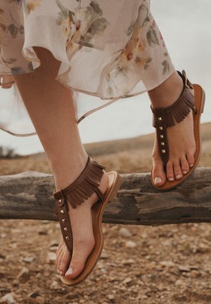 Voeten in bruine sandalen met franjes rustend op een houten balk, met een bloemenachtige transparante jurk die erboven golft.