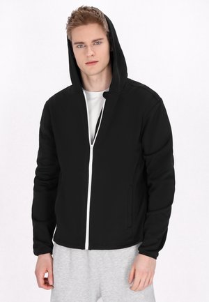 Zwarte capuchonjack met een volledige witte rits, zacht materiaal, elastische manchetten en twee zijzakken, gedragen over een wit shirt en grijze broek.