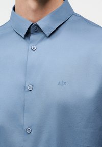 Κοντινό πλάνο ενός ανοιχτό μπλε πουκάμισου με γιακά και κεντημένο λογότυπο "A|X" στο στήθος.