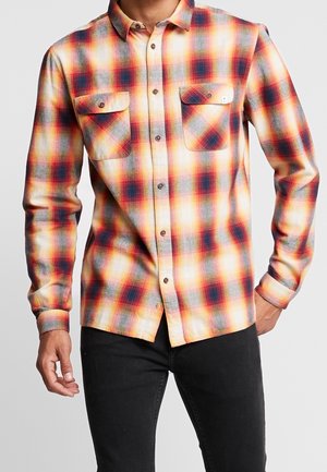 Camisa de franela a cuadros con tonos rojos, naranjas y grises, con un cuello camisa abotonado y dos bolsillos en el pecho. Textura suave y diseño ajustado.