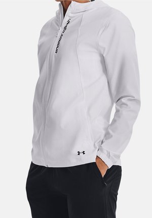 Person iført en hvid Under Armour hættetrøje med lynlås og hætte, sorte bukser og med den ene hånd i lommen, mod en ensfarvet baggrund.