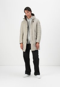 Beige polstret frakke med hætte, der har lynlås og knaplukning, sidelommer og et emblem på venstre ærme. Worn med sorte bukser og hvide sko.