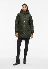 Quilted zaļš parka mētelis ar kapuci, spiedpogu aizdari un sānu kabatām. Valkāts virs melnajām šaurajām džinsām un melnajām zābakiem ar papēžiem.