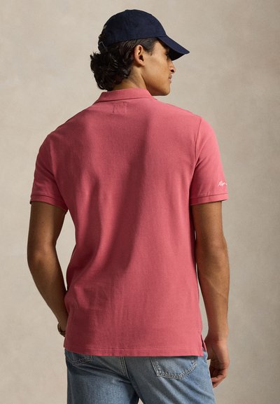 Άνδρας με σκούφο σκούρου χρώματος, ροζ κοντομάνικη πόλο μπλούζα και ανοιχτό μπλε τζιν, με την πλάτη γυρισμένη προς απλό φόντο.