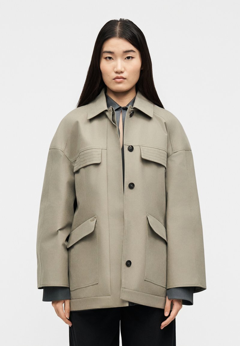 Νεαρή γυναίκα με μακριά σκούρα μαλλιά που φοράει μια υπερμεγέθη μπεζ ζακέτα με μεγάλες τσέπες και μαύρα κουμπιά, στέκεται μπροστά σε απλό φόντο.