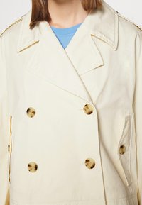 Manteau crème croisé à doubles rangées de boutons en écaille de tortue, porté par-dessus une chemise bleue, montrant le col et les détails du haut du torse.