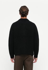 Mand med kort brunt hår iført en sort struktureret sweater og brune bukser, vendt væk mod en ensfarvet hvid baggrund.