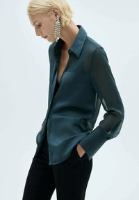Satin teal shirt with a button-down collar, sheer long sleeves, and a relaxed fit. Paired with dark pants, featuring side pockets.