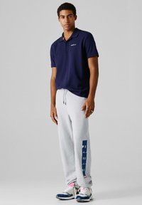 Jonge man staand in een marineblauw poloshirt, lichtgrijze joggingbroek met grote blauwe "REBEL" tekst en witte sneakers tegen een effen grijze achtergrond.