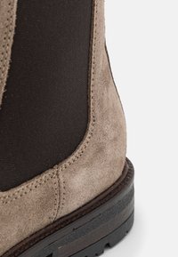 Bottine en daim marron avec panneau élastique noir, présentant des détails de couture, une semelle en caoutchouc texturée et un talon bas empilé.