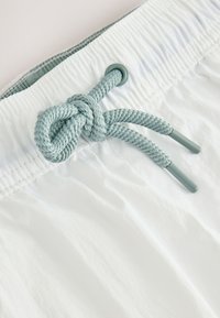 Hvide badeshorts med en tekstureret overflade, elastik i taljen og en lyseblå snor bundet i en knude. Fremhævet med metalliske ender.