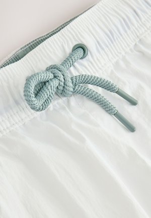 Fehér fürdőnadrág texturált felülettel, rugalmas derékrésszel és világoskék zsinórral, amely csomóra van kötve. Fém díszítővégekkel ellátva.
