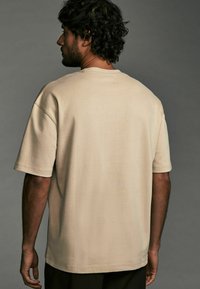 Beige bomulds t-shirt med en afslappet pasform, rund hals og korte ærmer. Teksturen er glat med synlige syninger på sømmene.