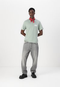 Lys grøn t-skjorte med en hvit logo, båret med grå jeans og svarte klumpete sko. Har et rødt bandana knyttet rundt halsen.