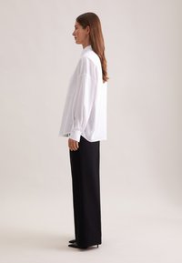 Camicia bianca oversize con colletto alto e maniche lunghe, abbinata a pantaloni neri a gamba dritta e décolleté neri a punta.