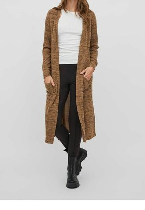 Femme portant un long cardigan marron ouvert avec des poches avant, un haut blanc, des leggings noirs et des bottes noires, debout devant un fond uni.