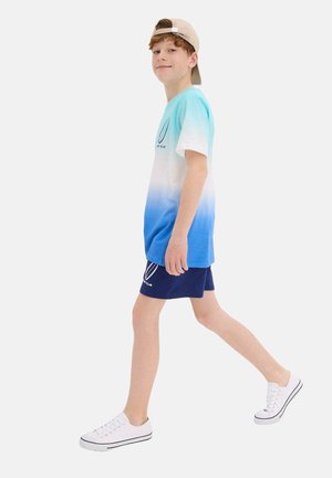 T-shirt à manches courtes dégradé bleu et blanc avec un logo, associé à un short navy et des baskets blanches, porté avec une casquette beige.