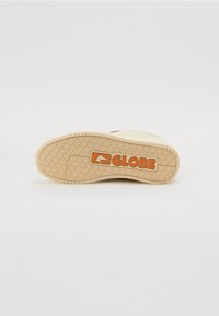 Rubberen zool met een patroon voor grip, in een tan kleur met een opgedrukte "GLOBE" logo in oranje. Bovenkant heeft een gladde, lichte afwerking.