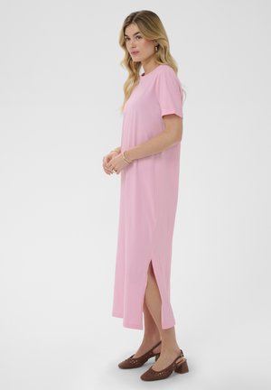 Donna con capelli biondi mossi che indossa un vestito midi rosa a maniche corte con spacco laterale e scarpe slingback intrecciate marroni con tacco.