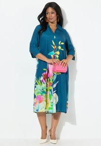 Vestido verde azulado con estampado floral en rosa, amarillo y verde; mangas tres cuartos; corte relajado; combinado con un bolso rosa y tacones blancos.