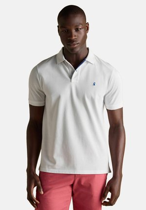 Weißes Poloshirt mit Kragen, das innen einen blauen Akzent hat, und einem kleinen blauen Logo auf der linken Brust. Kurze Ärmel und eine weiche Stofftextur.