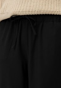 Un pantalon noir présente une texture lisse et une taille élastique avec un cordon de serrage noir. Un pull en tricot beige texturé est visible au-dessus.