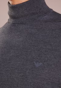 Mörkgrå turtlenecktröja i ribbad stickad kvalitet, med en subtil logotyp broderad i en ljusare nyans nära halsringningen.