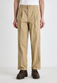 Khaki pants with pleats, mid-rise fit, and a flat front. Made from a durable fabric, paired with brown lace-up boots.