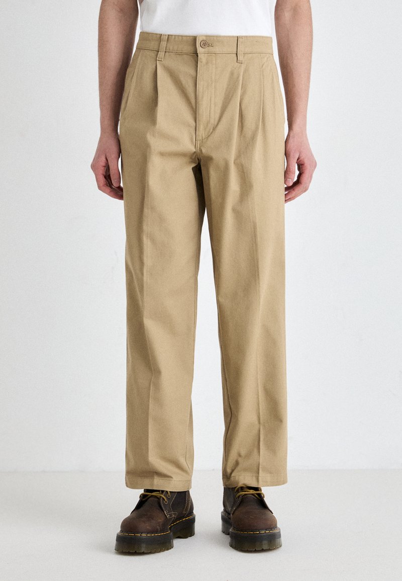 Khaki pants with pleats, mid-rise fit, and a flat front. Made from a durable fabric, paired with brown lace-up boots.