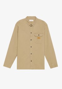 Camisa de manga larga color beige hecha de algodón, con cuello tipo botón, botones negros, un bolsillo en el pecho y una etiqueta de marca en el bolsillo.