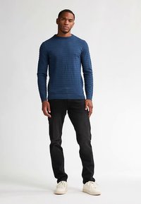 Tmavě modrý svetr s dlouhým rukávem a žebrovanou strukturou, kulatý výstřih, kombinovaný s černými kalhotami a krémovými teniskami, stojící před světle zbarveným pozadím.