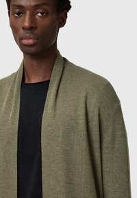Olivgrüner, offener Cardigan mit gerippter Textur und langen Ärmeln, getragen über einem schwarzen Shirt. Einfaches Design, ohne Verschlüsse oder Muster.