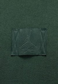 Ciemnozielona tkanina o dzianym wzorze; zawiera teksturowaną łatkę z logo Jumpman i napisem „FLY” wyszytym ciemnozieloną nicią.