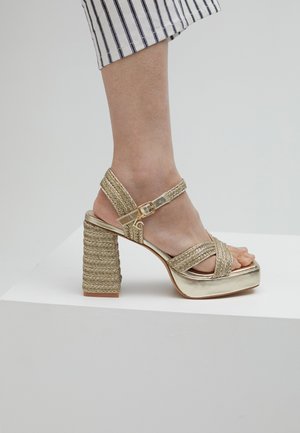 Gold platform sandals with woven texture. Strappy design featuring a buckle, high textured block heel, and rounded toe.